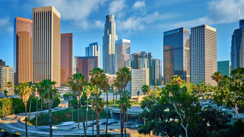 A vibrant cityscape of downtown Los Angeles, featuring tall skyscrapers and palm trees under a clear blue sky.