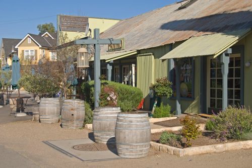 Quaint street scene featuring rustic buildings, wooden barrels, and greenery under a clear blue sky.
