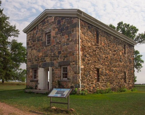 A historic stone building with barred windows, surrounded by grass and trees, featuring an informational sign in front.