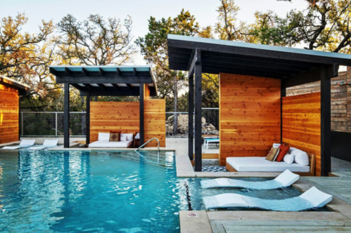 A serene pool area with wooden cabanas, lounge chairs, and lush greenery in the background.