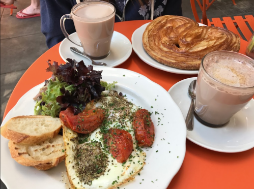 A plate with eggs, tomatoes, salad, and bread, alongside two cups of hot chocolate and a pastry on an orange table.