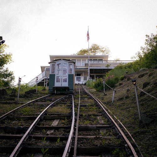 A green funicular car ascends a steep track towards a house, with an American flag visible in the background.