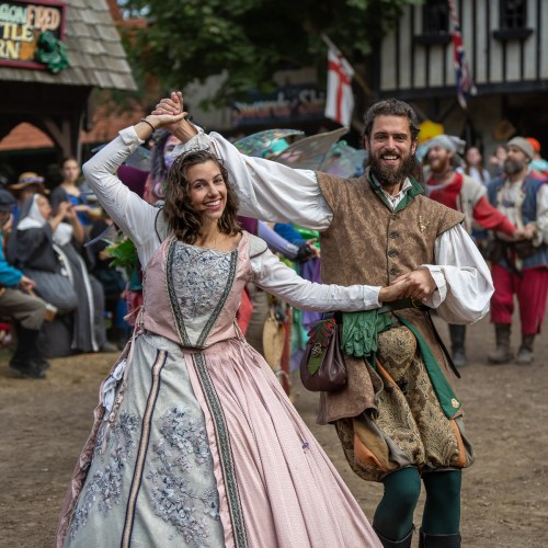 A man and woman in medieval costumes dance joyfully at a lively outdoor festival, surrounded by colorful performers.