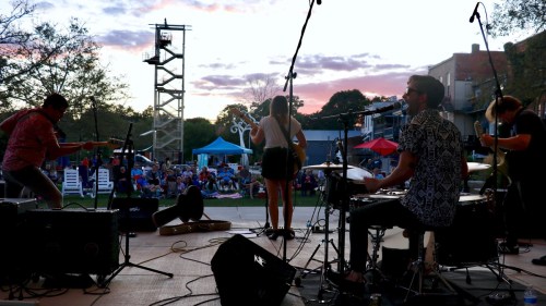 A band performs on stage at sunset, with an audience enjoying the show in the background.