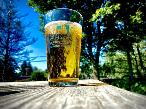 A glass of beer sits on a wooden table, surrounded by trees and a clear blue sky.