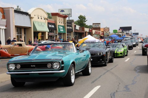 A parade of classic cars, including a turquoise convertible, drives down a street lined with shops and spectators.