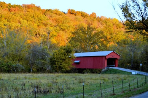 A red covered bridge surrounded by vibrant autumn foliage and a grassy field under a clear blue sky.
