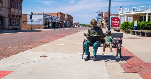 A bronze statue of an elderly man reading to a child on a bench in a quiet, sunny town street.