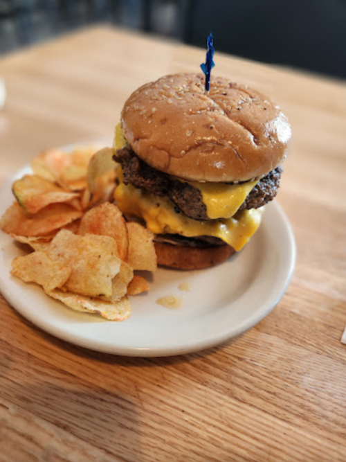 A juicy cheeseburger with a blue toothpick, served with a side of potato chips on a white plate.