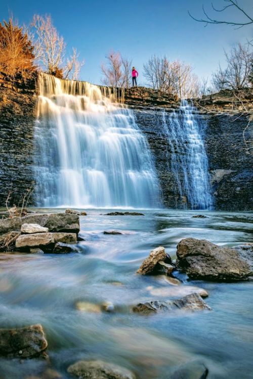 A waterfall cascades over rocky cliffs, with a person standing above, surrounded by trees and a clear blue sky.