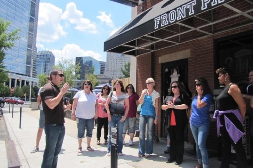 A group of people gathered outside a building, listening to a speaker under a sunny sky with city buildings in the background.