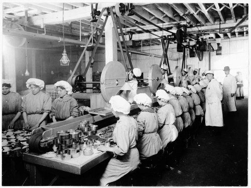 Black and white photo of workers in a factory, assembling products on a conveyor belt in a large industrial space.