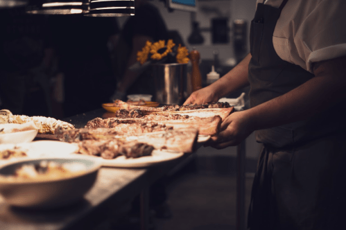 A chef prepares various cuts of meat on a countertop, with bowls of food and flowers in the background.