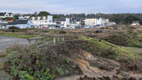 A coastal town with colorful buildings, grassy areas, and a cloudy sky in the background.