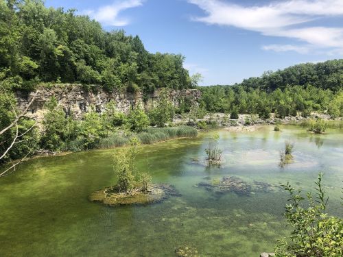 A serene landscape featuring a green pond surrounded by rocky cliffs and lush trees under a clear blue sky.