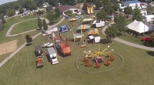 Aerial view of a fairground with rides, tents, and food trucks set in a green park.
