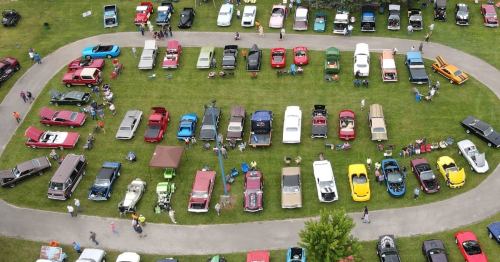 Aerial view of a car show featuring a variety of classic cars parked on a grassy area with spectators.
