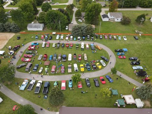 Aerial view of a car show featuring a variety of classic cars arranged in a circular layout on a grassy area.