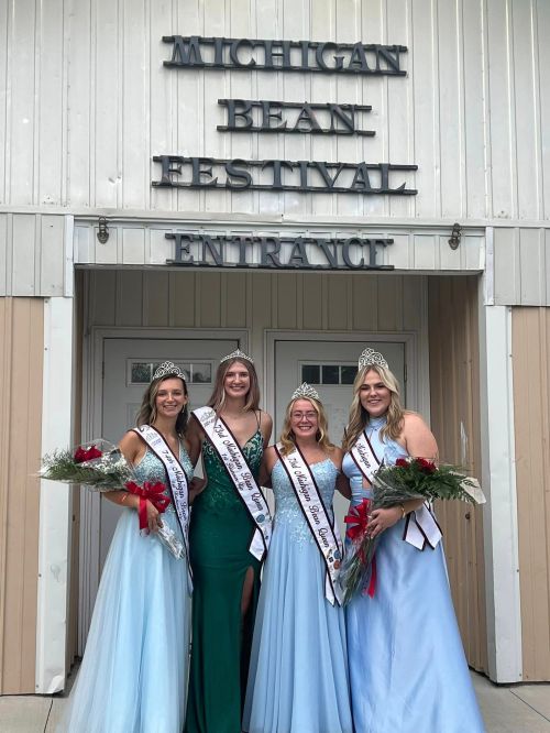 Four pageant winners in formal dresses and tiaras stand in front of the Michigan Bean Festival entrance, holding bouquets.