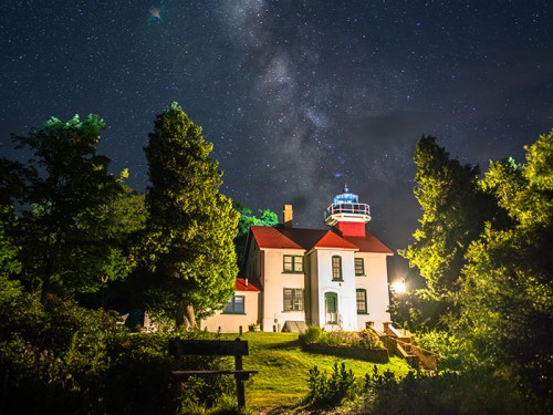 A lighthouse at night, surrounded by trees, under a starry sky with the Milky Way visible above.