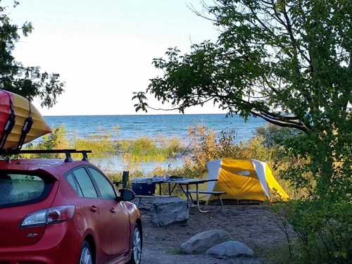 A red car with kayaks on top parked near a campsite by the water, featuring a yellow tent and picnic table.