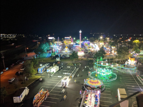Aerial view of a vibrant night fair with colorful lights, rides, and parked vehicles in a spacious lot.
