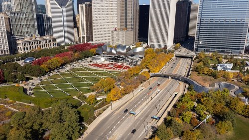 Aerial view of a city park with colorful trees, modern buildings, and a road running alongside the green space.