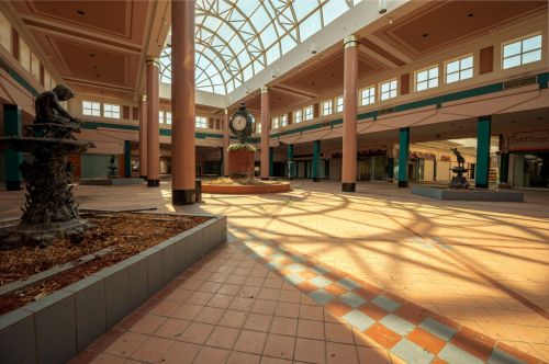 Abandoned indoor space with a glass ceiling, empty shops, and decorative fountains casting shadows on the tiled floor.