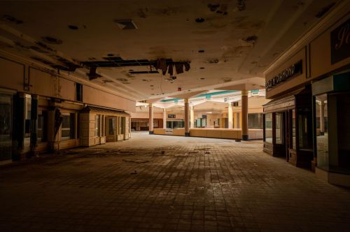 Abandoned shopping mall interior with damaged ceilings and empty storefronts, showcasing neglect and decay.