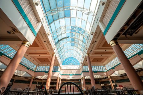 A spacious indoor mall with a glass ceiling, featuring tall columns and a bright blue sky visible above.