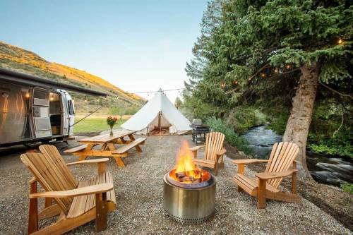 A cozy campsite with a fire pit, wooden chairs, a tent, and an Airstream trailer by a stream and mountains.