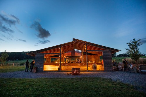 A rustic wooden building illuminated at dusk, surrounded by greenery and people enjoying the outdoor space.