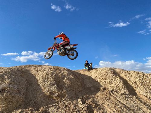 A motocross rider jumps over a dirt mound while another person watches from the side against a blue sky.