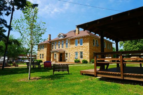 Historic stone building with a porch, surrounded by green grass and trees under a clear blue sky.