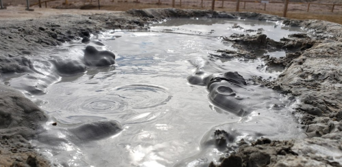A bubbling mud pool with grayish mud and water, surrounded by dry terrain.