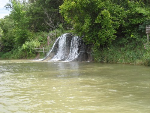 A small waterfall cascades over rocks into a calm, greenish river, surrounded by lush trees and vegetation.