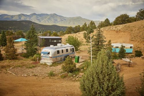 A vintage Airstream trailer surrounded by trees and mountains, with a blue umbrella and another trailer in the background.