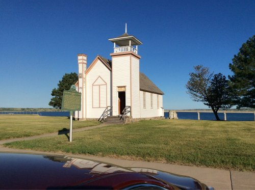 A small white church with a bell tower, set against a clear blue sky and near a body of water. Green grass in the foreground.