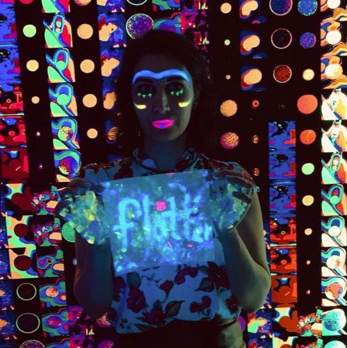 A person with neon face paint holds a glowing sign that says "flatt" against a vibrant, colorful background.