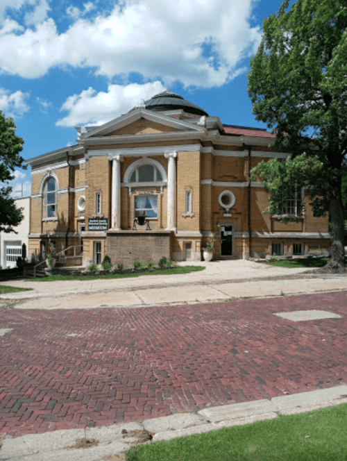 Historic brick building with columns, large windows, and a dome, surrounded by greenery and a brick-paved street.