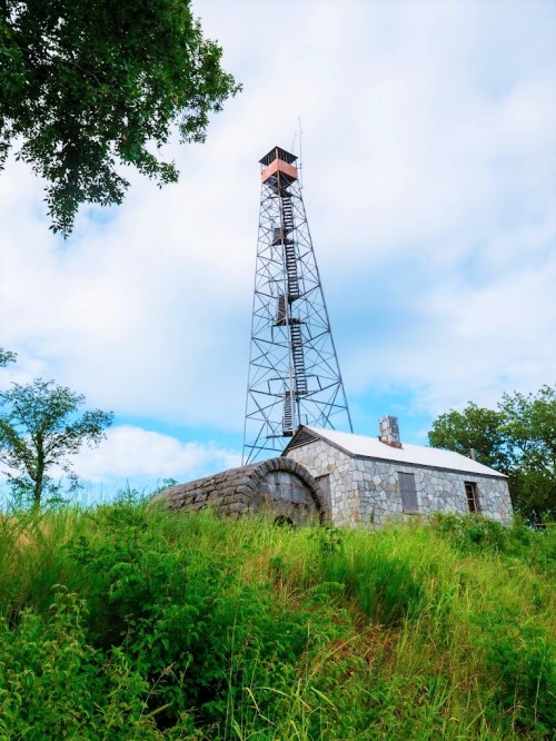 A tall metal tower stands beside a stone building, surrounded by lush green grass and trees under a cloudy sky.