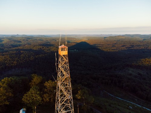 A tall fire lookout tower stands on a mountain, surrounded by lush green forests and rolling hills under a clear sky.