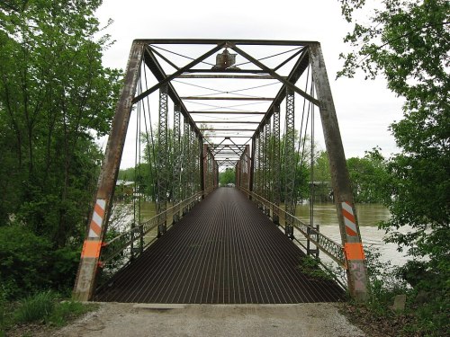 A weathered metal bridge spans a river, surrounded by lush greenery and a cloudy sky.