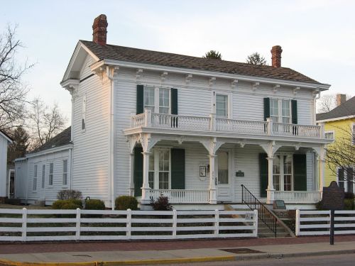 A white Victorian-style house with green shutters, a porch, and a picket fence, set against a clear sky.