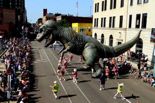 A large dinosaur balloon floats above a parade, with crowds watching and participants in costumes walking along the street.