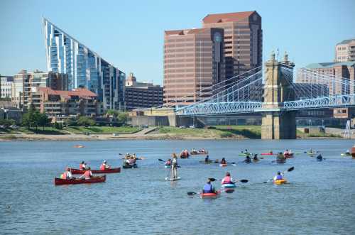 A vibrant scene of kayakers on a river with a city skyline and a bridge in the background on a sunny day.