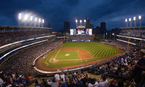 A panoramic view of a packed baseball stadium under a cloudy sky, with bright lights illuminating the field.