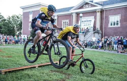 Two cyclists jump over a ramp, one on a mountain bike and the other on a smaller bike, with a crowd watching in the background.