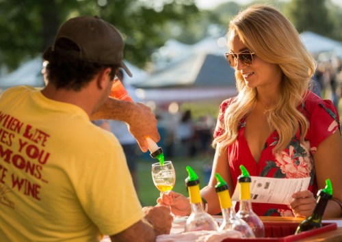 A woman in sunglasses watches as a vendor pours a drink at an outdoor event, surrounded by tents and greenery.