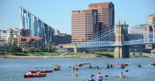A vibrant scene of kayakers on a river with a city skyline and a blue bridge in the background.